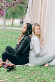 Two diverse women meditate in peaceful outdoor setting, back to back.