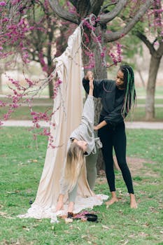 Two women practicing aerial yoga with a tree and hammock outdoors.