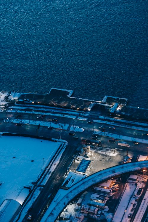 Free Drone view of snowy seafront with structures and cars riding on road near rippling sea in evening time in city Stock Photo