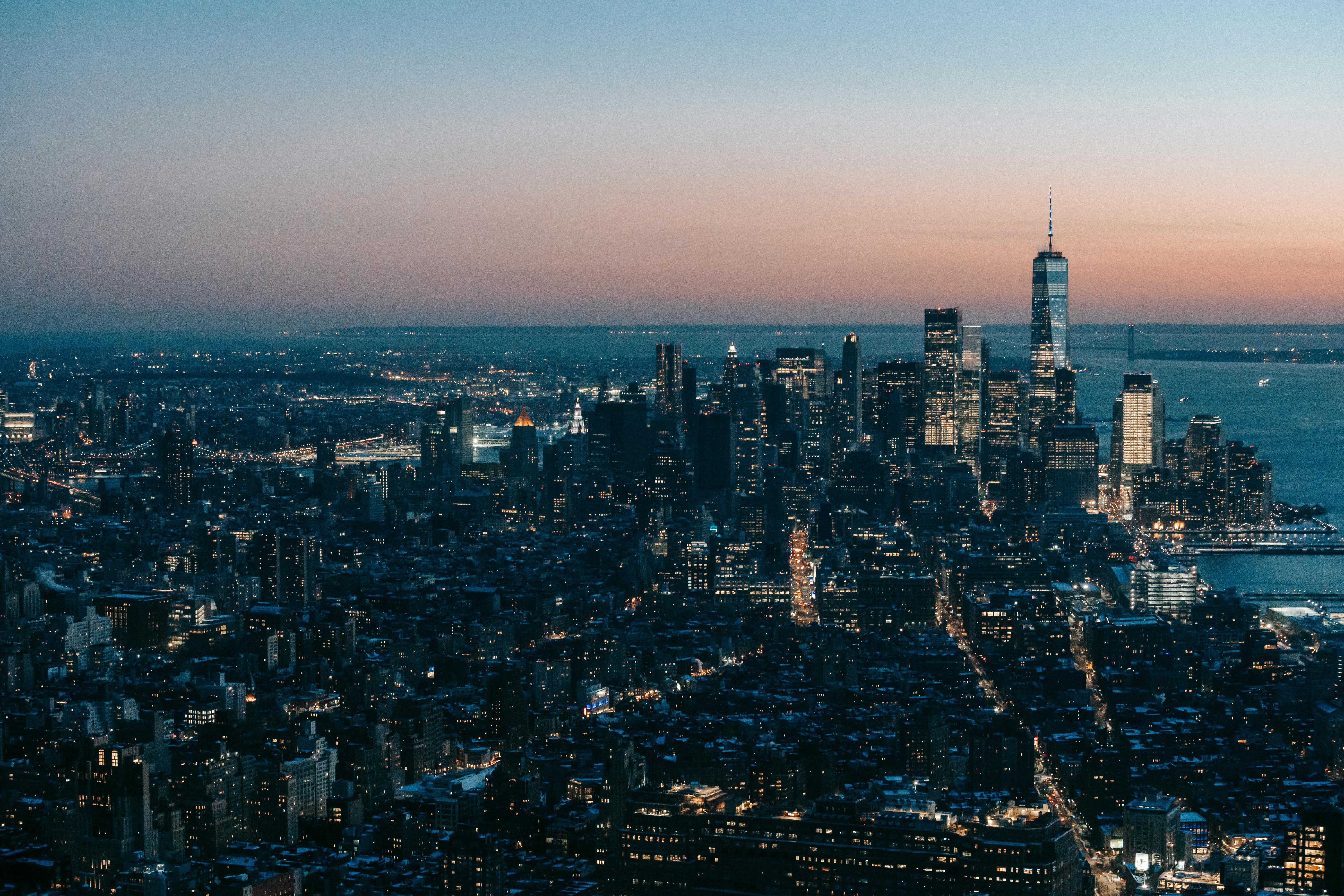 Modern multistory buildings and skyscrapers with glowing lights located on streets in megapolis near calm sea against cloudless sky in evening time