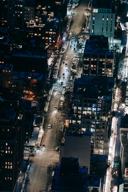 Drone view of road with cars riding between modern multistory buildings with glowing lights located on street of city at night time