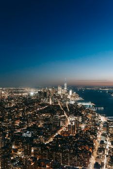 Modern buildings with glowing lights located on streets of contemporary megapolis near calm sea against cloudless sky at night time