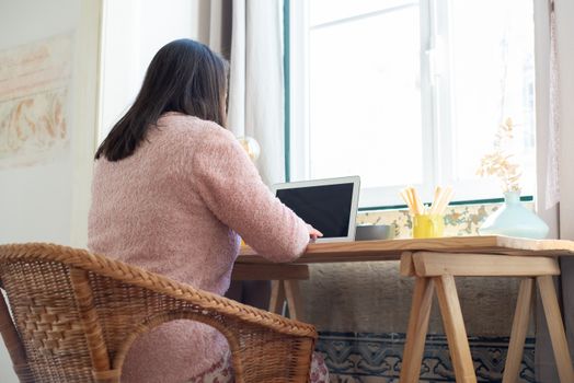 A businesswoman works at a wooden desk near a window, using a laptop in a cozy home setting.