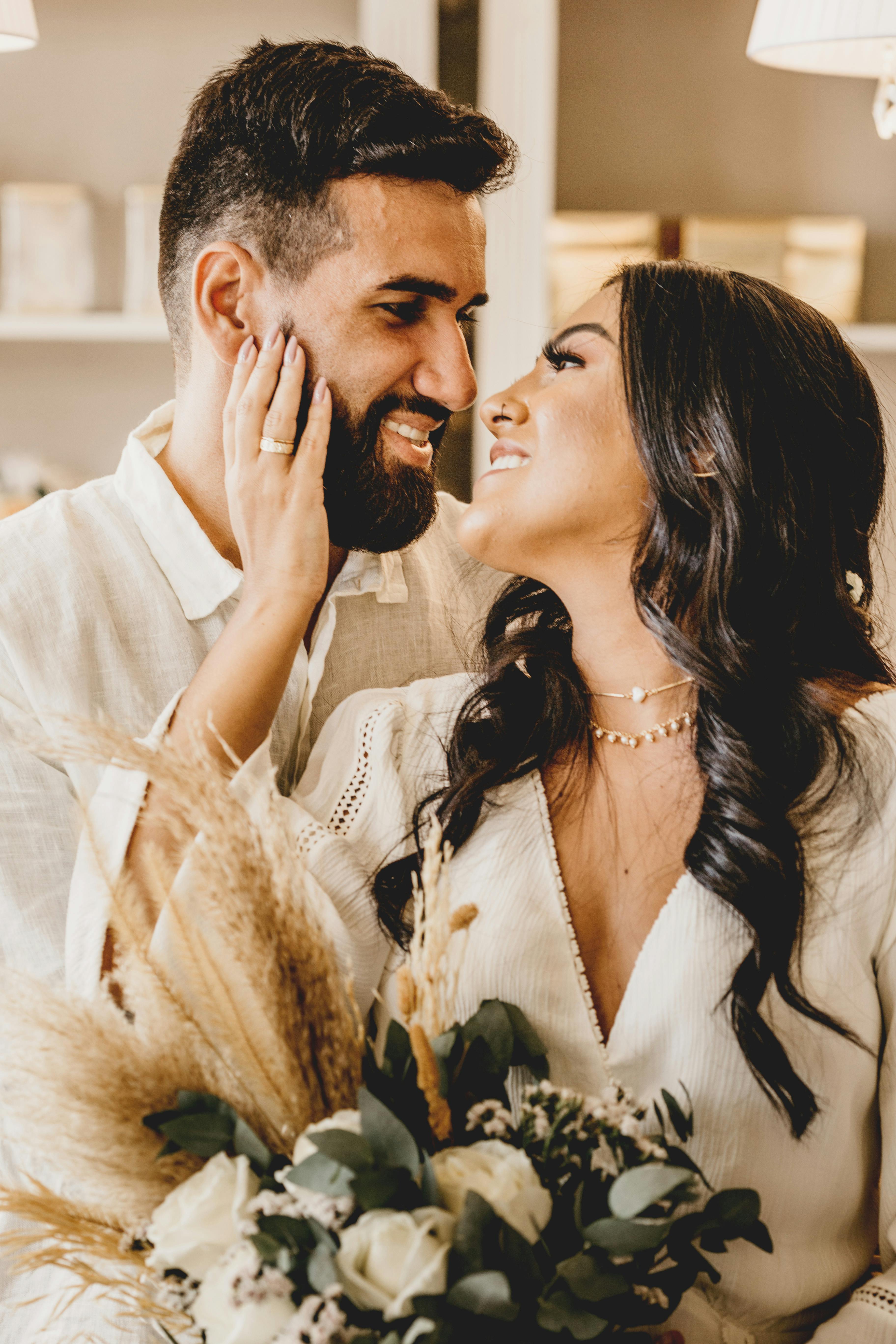 cheerful ethnic newlywed couple with rose bouquet embracing in room