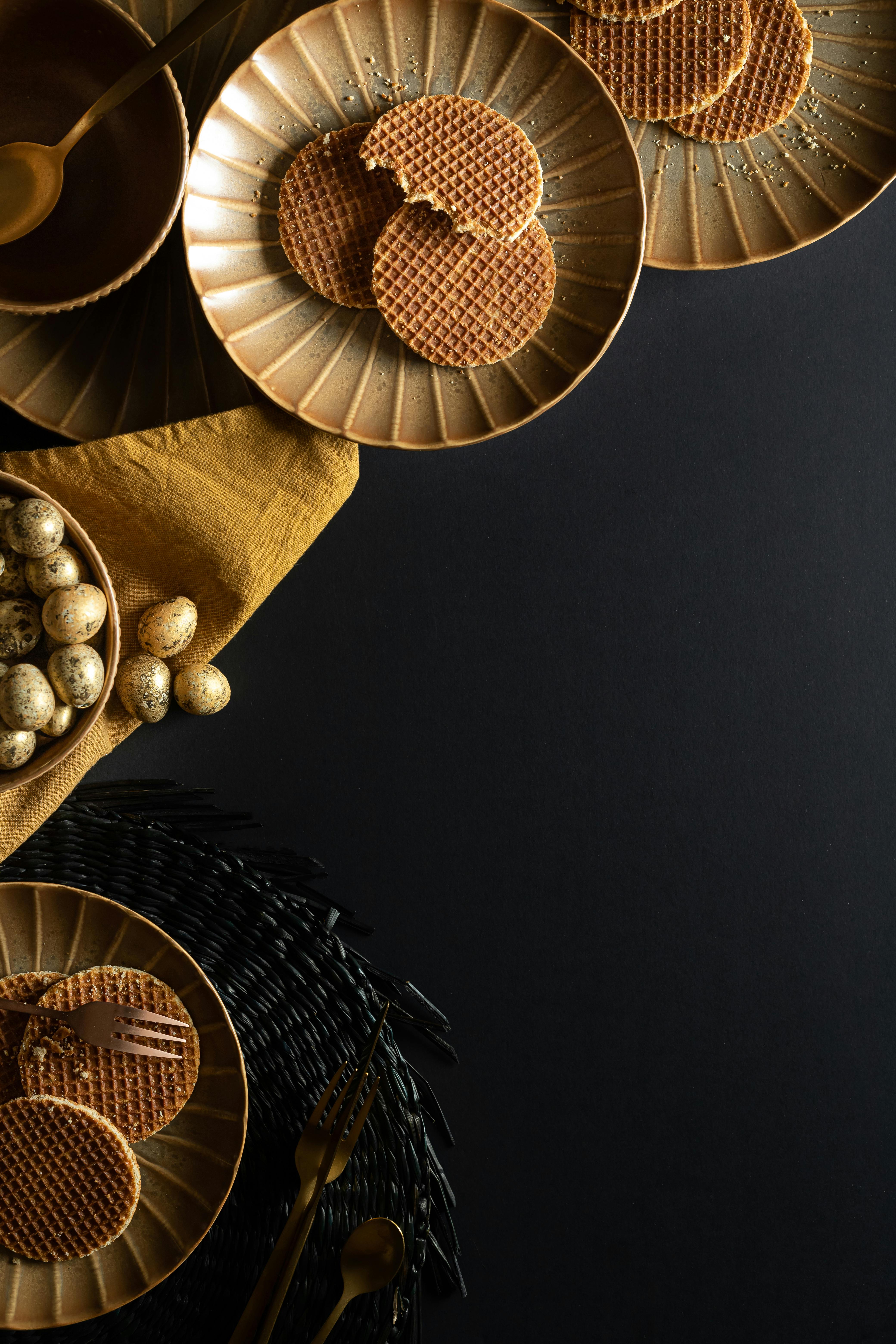 Stylish top view of ceramic plates with waffles, quail eggs, and utensils on a black background.