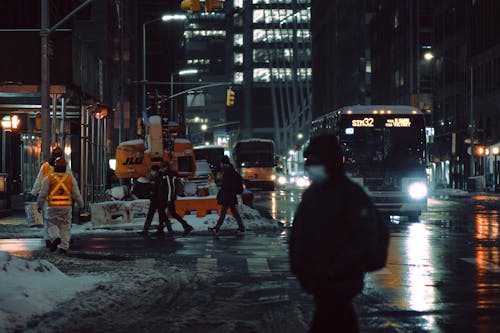 People in medical masks strolling on crosswalk near roadway with glowing buses on dark street during winter evening in city