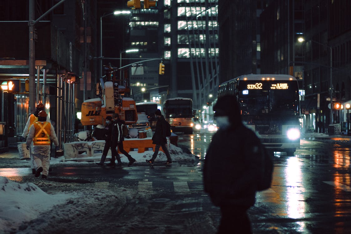 Free People in medical masks strolling on crosswalk near roadway with glowing buses on dark street during winter evening in city Stock Photo