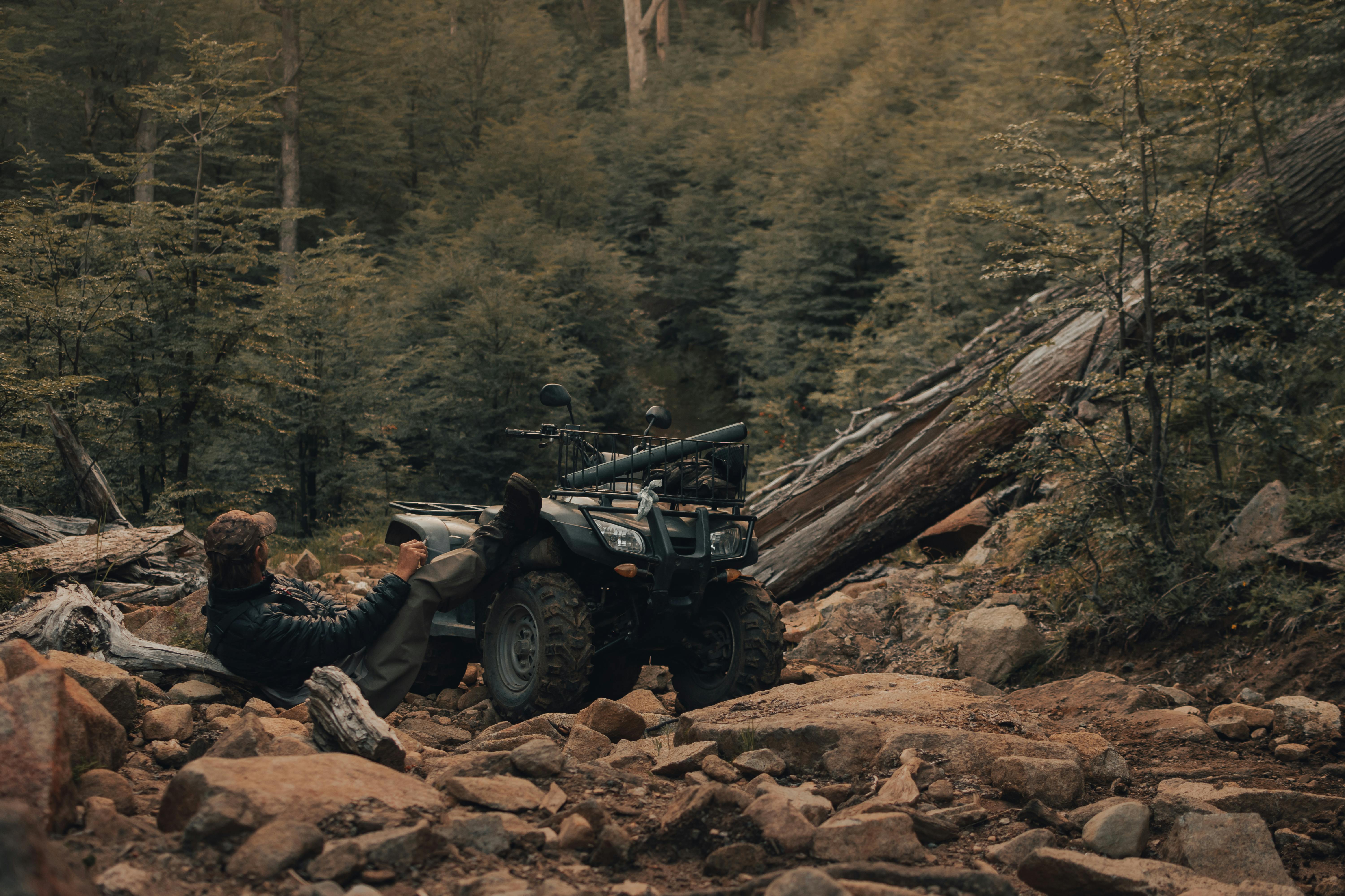 A man relaxing beside an ATV in a dense forest setting, surrounded by rocks and trees.