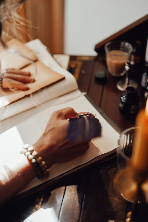 Free Crop anonymous female writer taking notes in copybook with feather at vintage table in sun ray at home Stock Photo