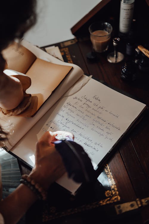 From above of crop unrecognizable female writer taking notes in copybook with feather at vintage table in sunbeam