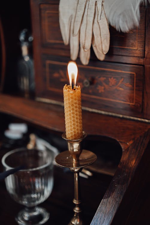 Burning candle placed on antique wooden writing cabinet with glass cup and writing supplies on blurred background in vintage store