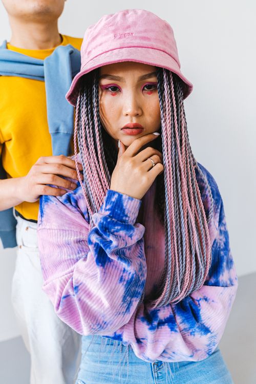 Free A fashionable Asian woman wearing a pink bucket hat and tie-dye sweatshirt, posing indoors. Stock Photo