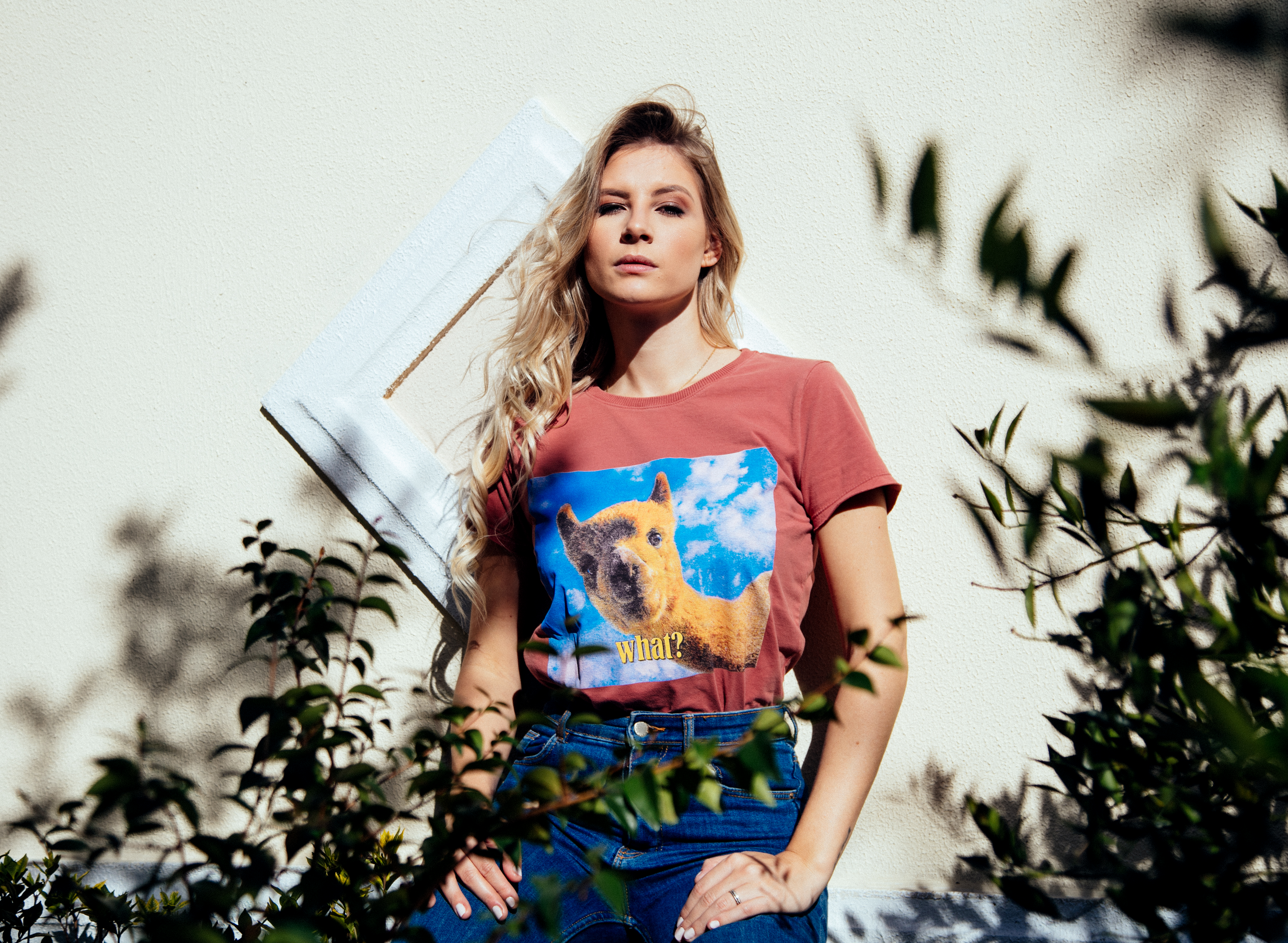 Stylish blonde woman in a graphic tee poses confidently in sunlight outdoors, surrounded by plants.