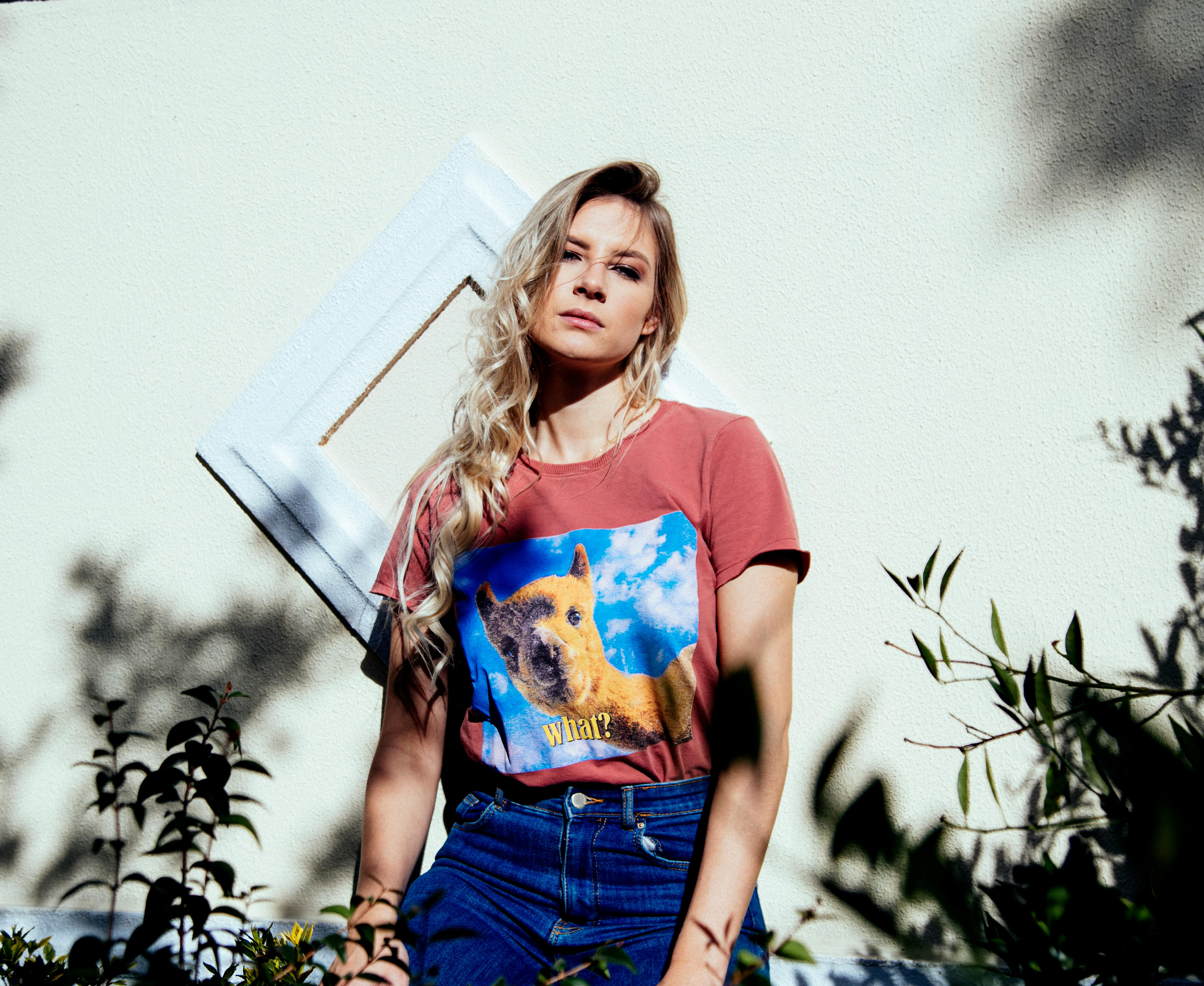 Blonde woman poses in a graphic tee, outdoors in Curitiba, casting dynamic shadows.