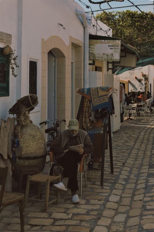 Free Elderly man leisurely reading on cobblestone street in charming outdoor setting. Stock Photo