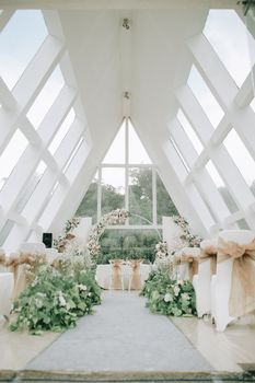 Interior of light gazebo with white chairs and arch decorated with flowers and plants for wedding ceremony near windows in daylight under bright sky near trees
