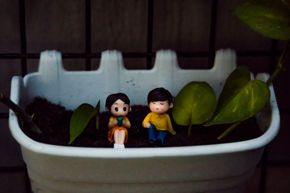Cute colorful figurines of boy and girl placed in white flowerpot with green leaves of plant near metal fence in garden