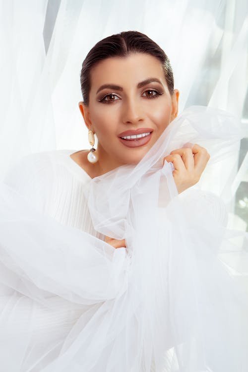 Charming female with makeup smiling and looking at camera while standing near window with white tulle in hands in room