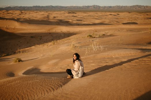 A woman sitting alone on sand dunes, enjoying the calm desert scenery under the warm sun.