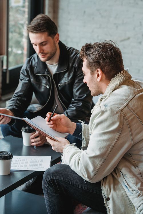 Free Casual male colleagues sitting together at table and taking notes while drinking coffee in cafe on daytime Stock Photo
