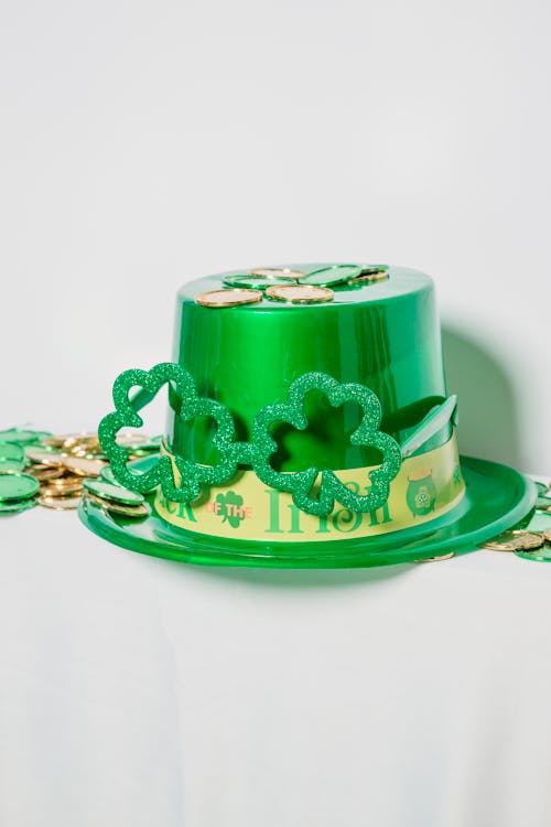 Shiny green hat with decorations and clover placed near heap of colorful coins on white background