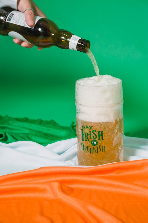 Crop anonymous person with ring pouring beer with froth from bottle into glass mug placed on Irish flag on green background