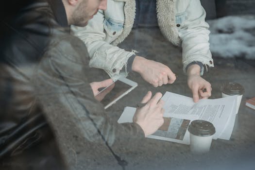 Through glass from above of crop male entrepreneurs sitting at table with takeaway coffee and reading contract while using tablet