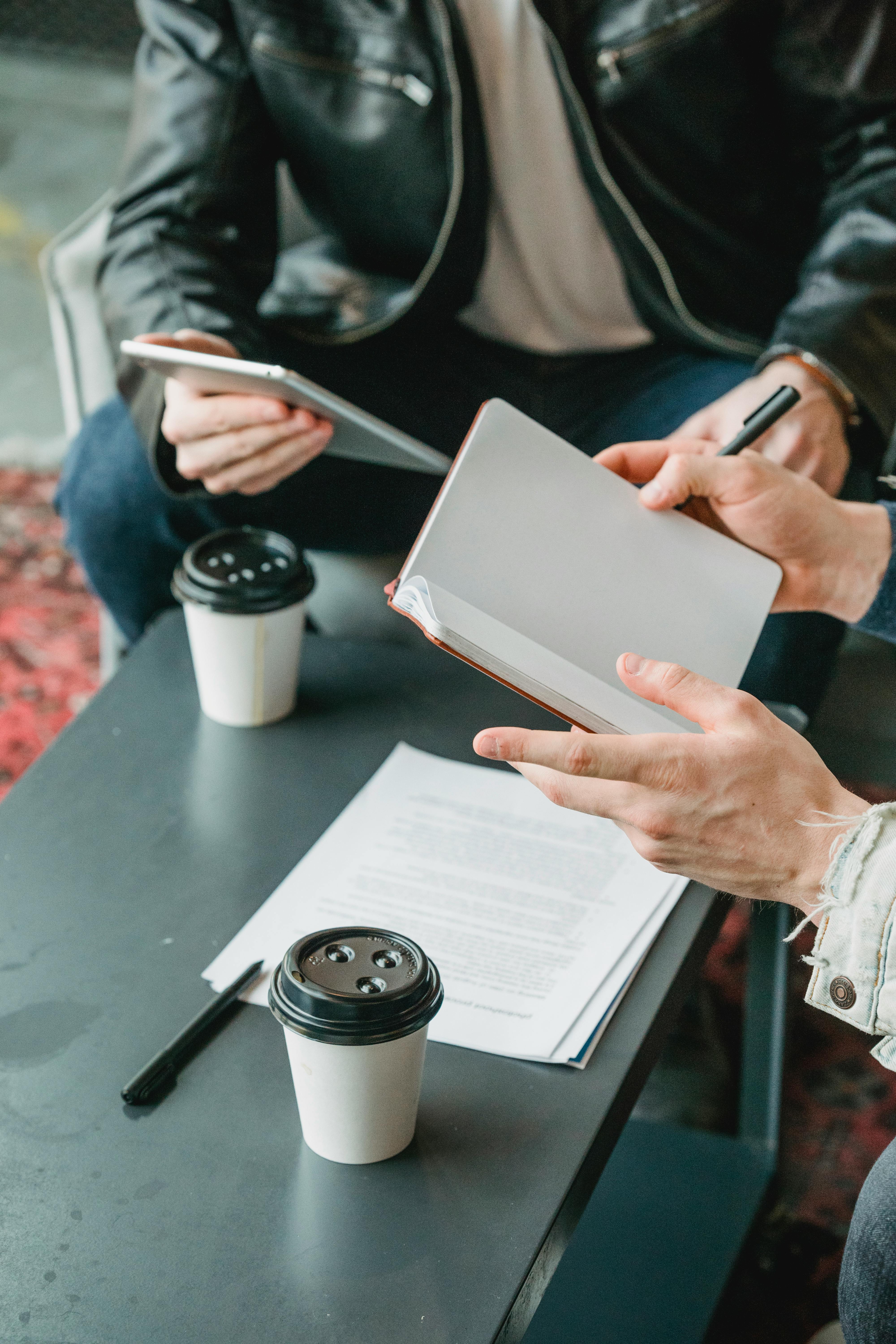 Free Two businessmen collaborating over notebooks and coffee in a casual meeting setting. Stock Photo