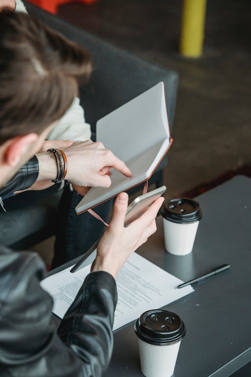 From above of crop anonymous male entrepreneur pointing in notepad of coworker while sitting at table with contract and coffee cups