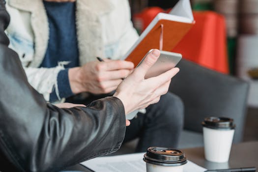 Crop anonymous man showing tablet to male partner sitting at table with notebook and coffee cups during work in cafe