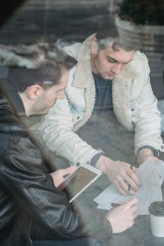 Through glass high angle of concentrated young men in casual clothes sitting at table and checking papers while working with tablet
