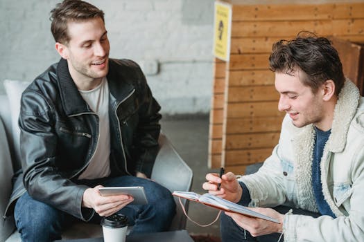 Two young men having a collaborative meeting, using a tablet and notepad in a casual indoor setting.