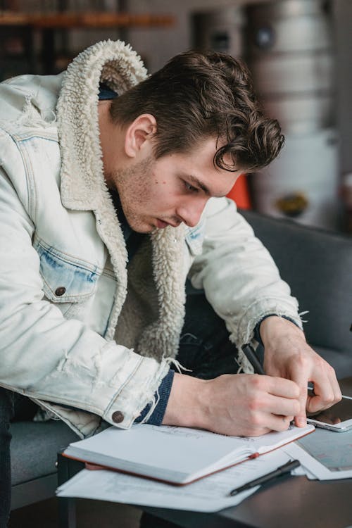 Focused male worker writing notes in notepad while sitting on sofa at table with tablet