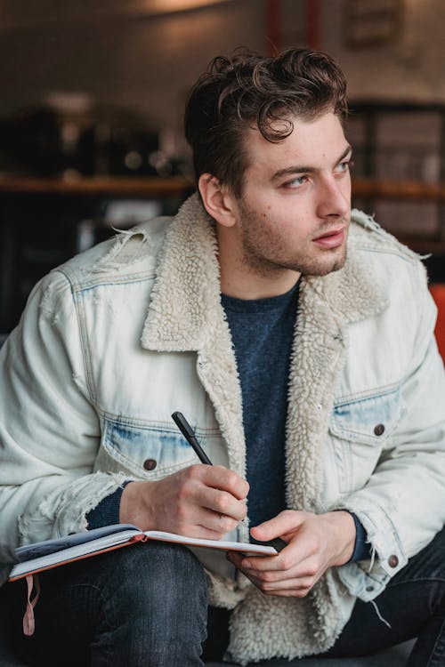 Serious male in casual clothes sitting alone and writing thoughts in notebook while looking away