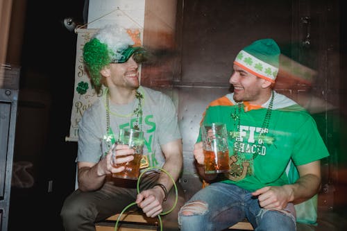 Through glass wall view of smiling male partners with mugs of beer looking at each other during Feast of Saint Patrick