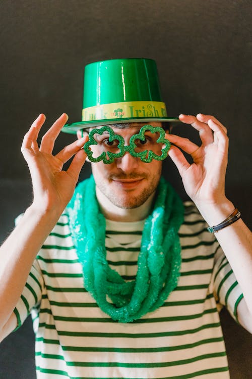 Male in shamrock hat with decorative eyeglasses looking at camera during Feast of Saint Patrick on gray background