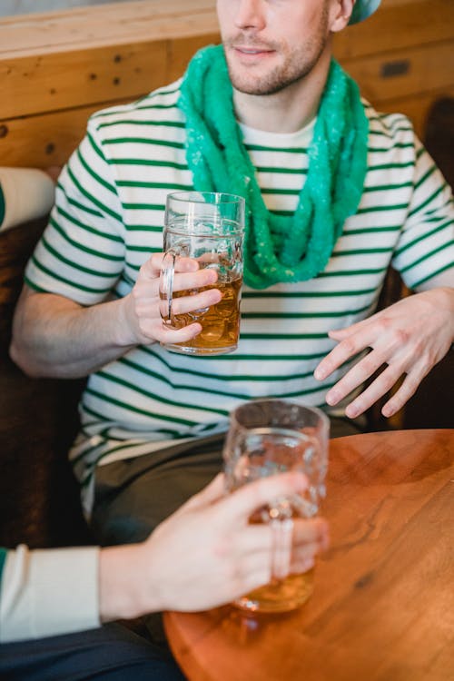 Crop anonymous male partners with jars of beer talking while sitting on bench at bar table