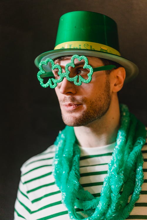 Contemplative male in green hat and eyeglasses with shamrock ornament looking away during Feast of Saint Patrick on dark background