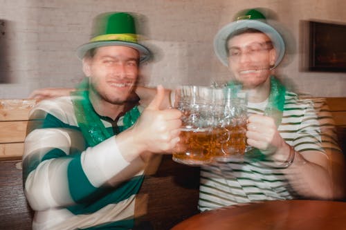 Content male partners in shamrock hats with jars of beer celebrating Feast of Saint Patrick at table in pub