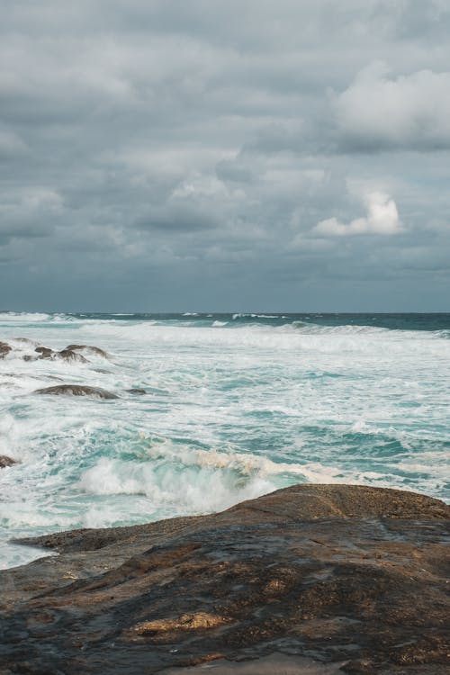 Scenery of azure sea with foamy waves rolling on wet rough seacoast beneath dull cloudy sky