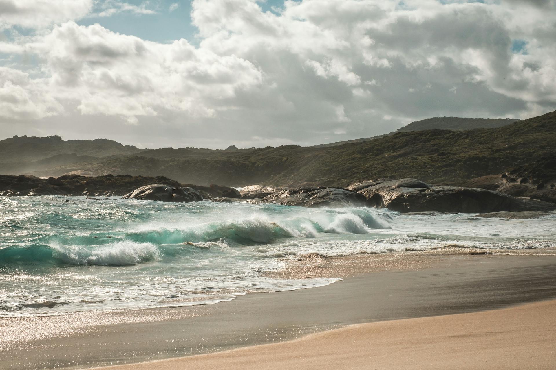 Scenery of azure foamy sea waving on sandy beach near rough grassy cliff on sunny day