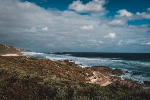 Free Majestic scenery of endless turquoise ocean with powerful foamy waves washing sandy coast and rough boulders near rocky mountain covered with green grass under cloudy blue sky Stock Photo