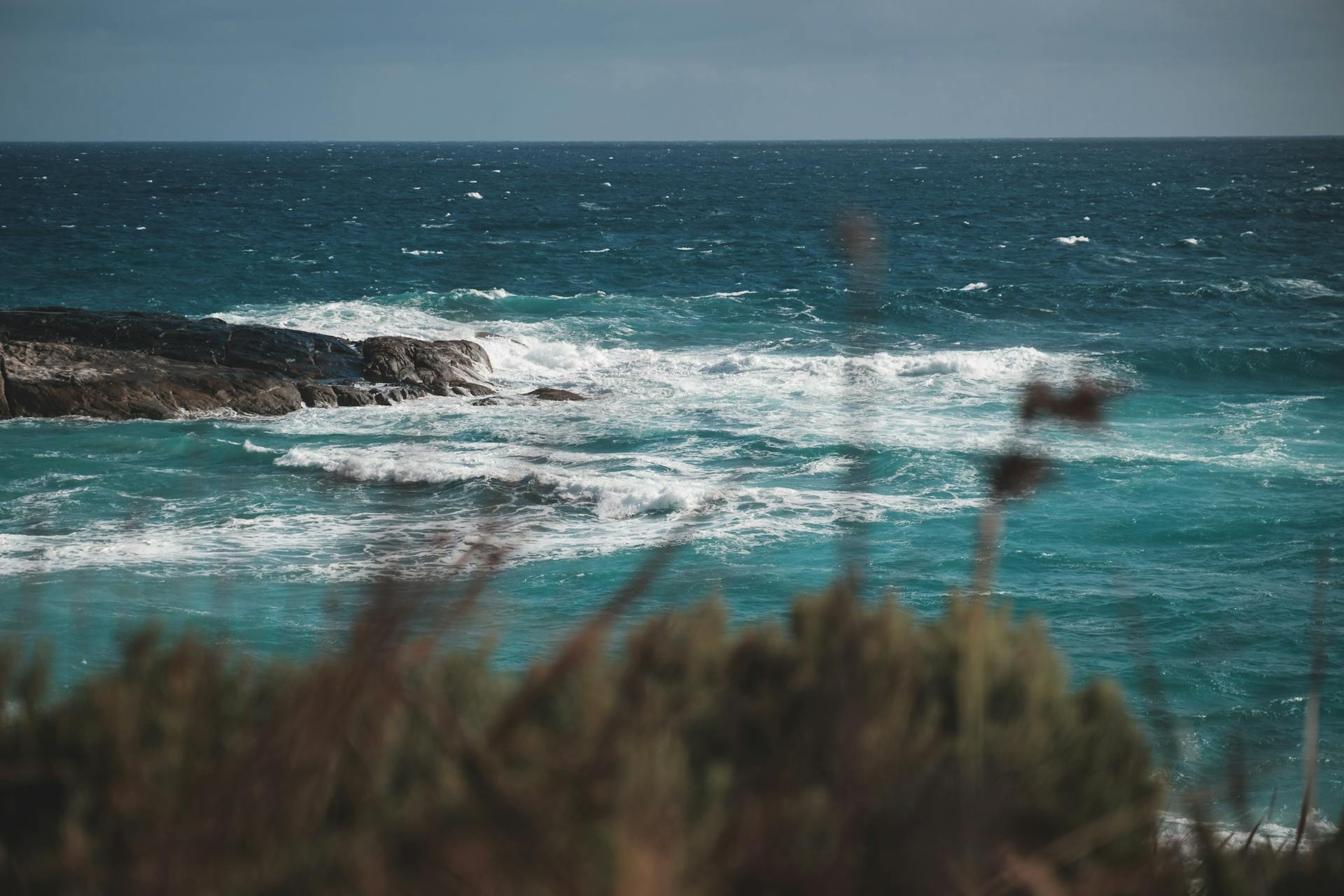 Scenic endless ocean with azure water and foamy waves splashing on rocky formation near beach surrounded by hill with lush green plants