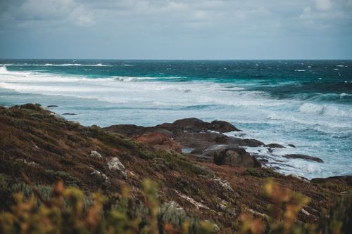 Free Rocky boulders washing by foamy waves of powerful ocean near mountain covered with grass and blooming plants against cloudy sky Stock Photo