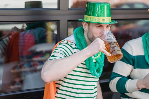 Young unshaven male millennial in casual clothes and green leprechaun hat drinking mug of cold beer and looking away during celebration pf St Patricks Day with crop anonymous friend