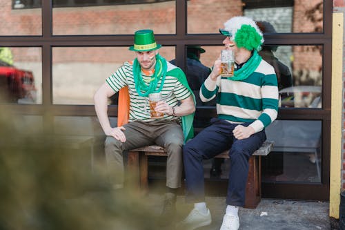 Happy young male friends in festive outfits drinking beer and communicating while relaxing on street bench during celebration of St Patricks Day