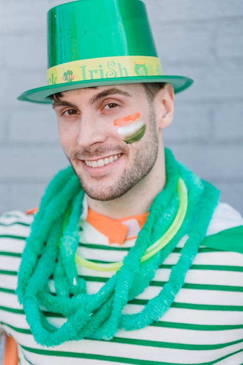 Delighted male in traditional hat and flag of Ireland painted on face looking at camera while standing on street in city