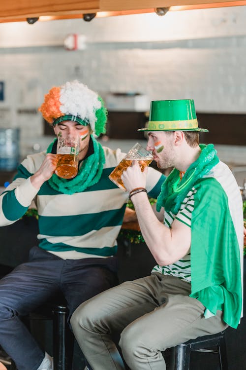 Free Male friends in traditional clothes and hats drinking beer while sitting on chairs at counter in cafe during Saint Patricks day Stock Photo