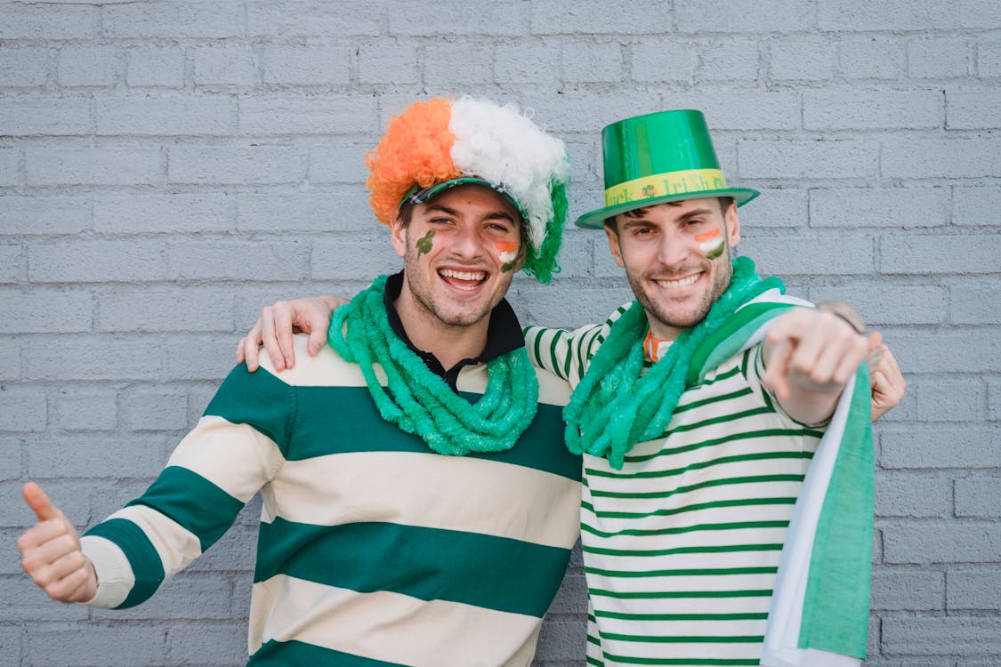Free Delighted male friends in traditional colorful hats and flags of Ireland painted on faces looking at camera during holiday on street Stock Photo