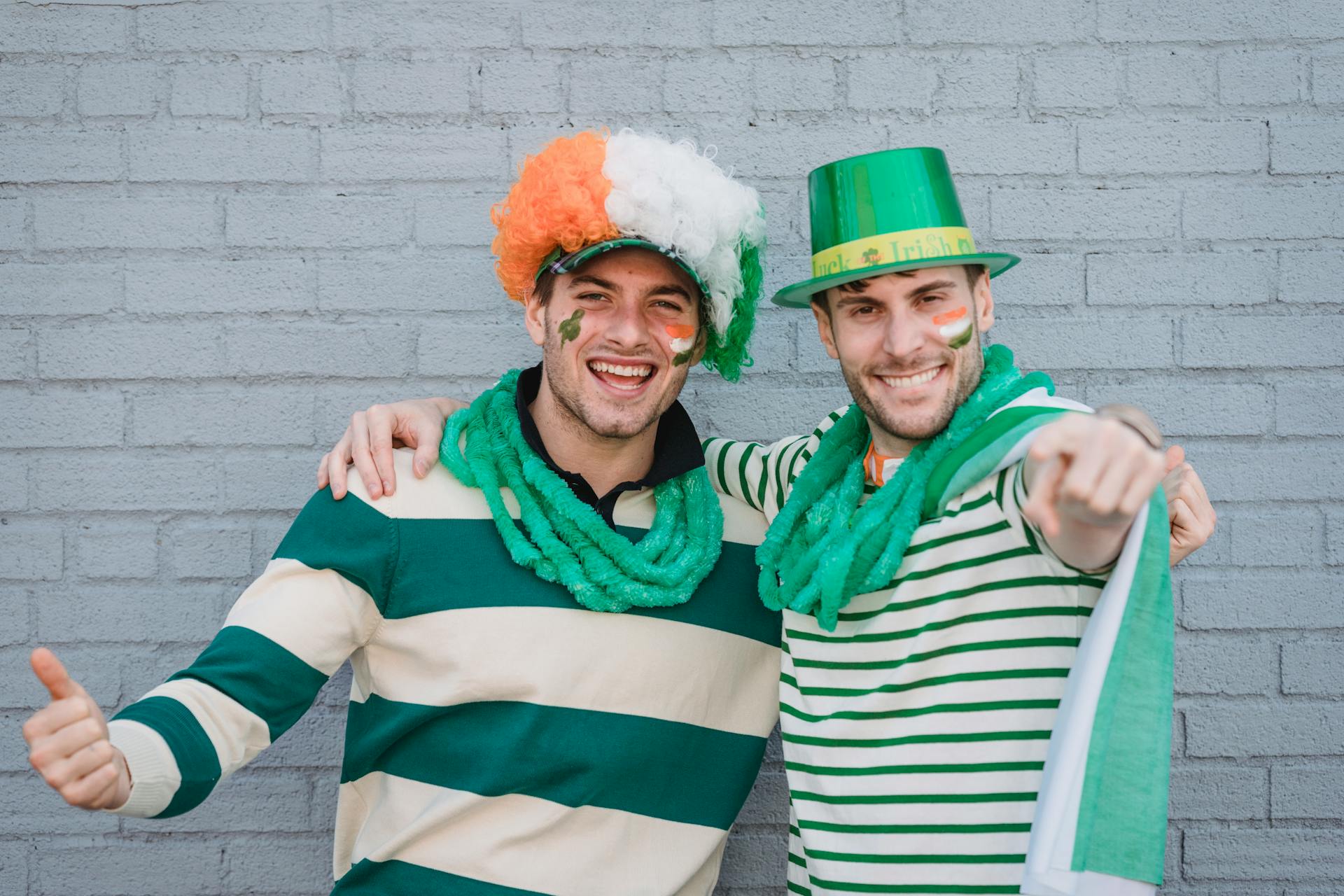 Delighted male friends in traditional colorful hats and flags of Ireland painted on faces looking at camera during holiday on street